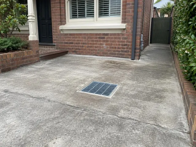 Clean outdoor drain grate set in dry concrete paving beside a brick house in Clovelly
